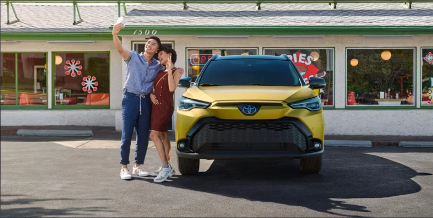 A man and a woman taking a selfie next to a yellow 2024 Toyota Corolla Cross Hybrid.