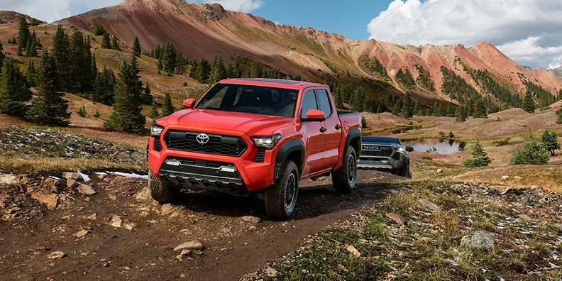 A red 2024 Toyota Tacoma off-roading down a dirt road with mountains in the background.