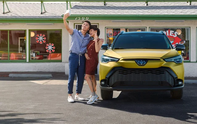 Two individuals taking a selfie in front of a yellow 2024 Toyota Corolla Cross.