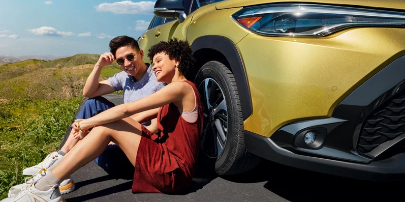 Two individuals sitting next to a yellow 2024 Corolla Cross.