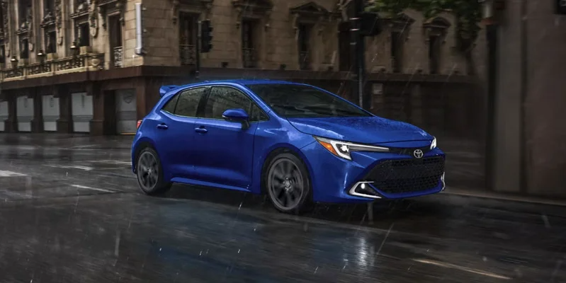 A photo of a blue 2026 Toyota Corolla Hatchback driving down the road in the rain.