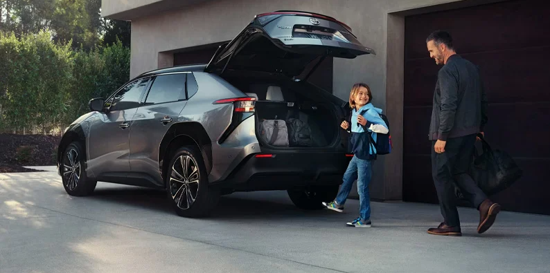 A man and child walking toward the back of a 2025 Toyota bZ4X parked in a driveway.