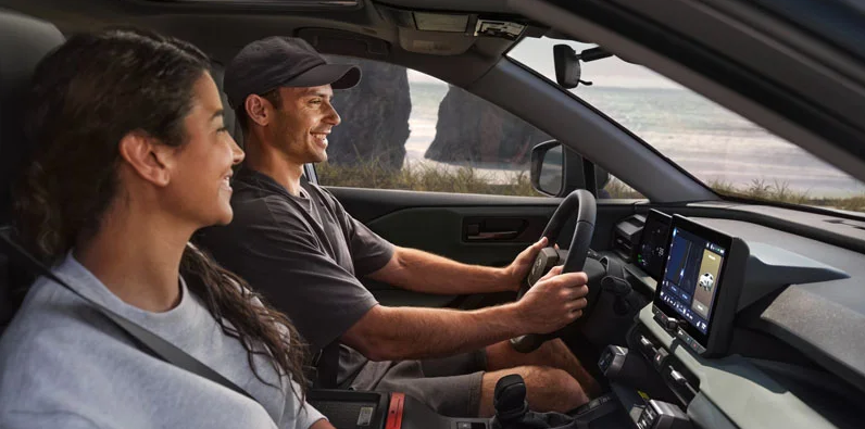 A man and woman driving down the road with an ocean outside the drivers window.