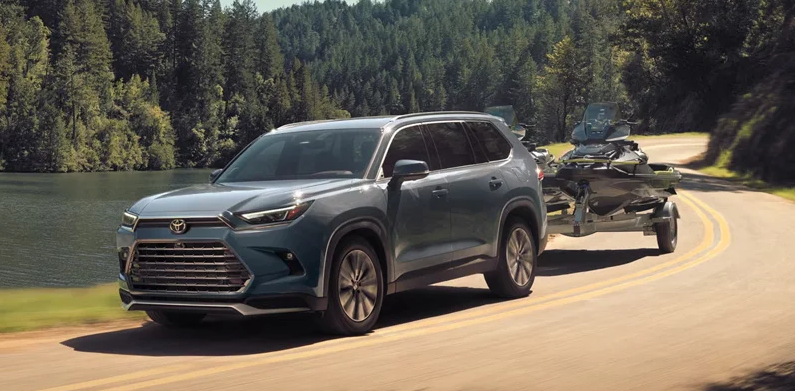 A 2026 Toyota Grand highlander towing a jet ski down a lake side road.