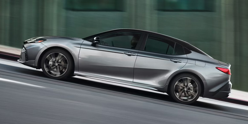 A sleek silver 2026 Toyota Camry driving up a city street, showcasing its dynamic design and modern aesthetic.