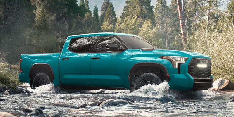 A bold 2026 Toyota Tundra in vibrant turquoise, navigating through a river in a rugged, outdoor setting.